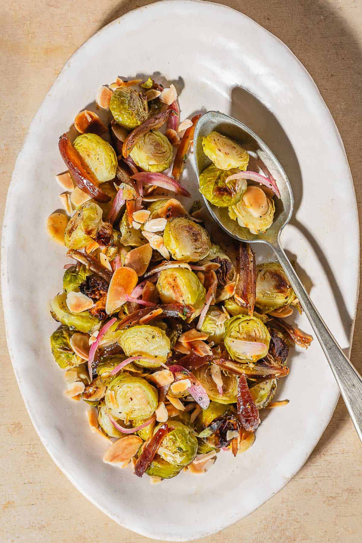 An overhead photo of roasted brussels sprouts with dates and almonds on a platter with a spoon.