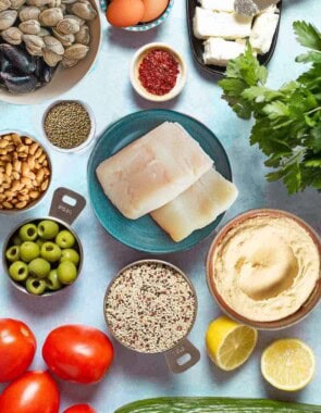 An overhead photo of several Mediterranean diet friend foods including eggs, fish, clams, mussels, feta cheese, aleppo pepper, lentils, quinoa, beans, olives, tomatoes, lemons, cucumber, hummus and parsley.