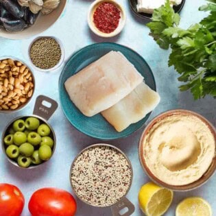 An overhead photo of several Mediterranean diet friend foods including eggs, fish, clams, mussels, feta cheese, aleppo pepper, lentils, quinoa, beans, olives, tomatoes, lemons, cucumber, hummus and parsley.