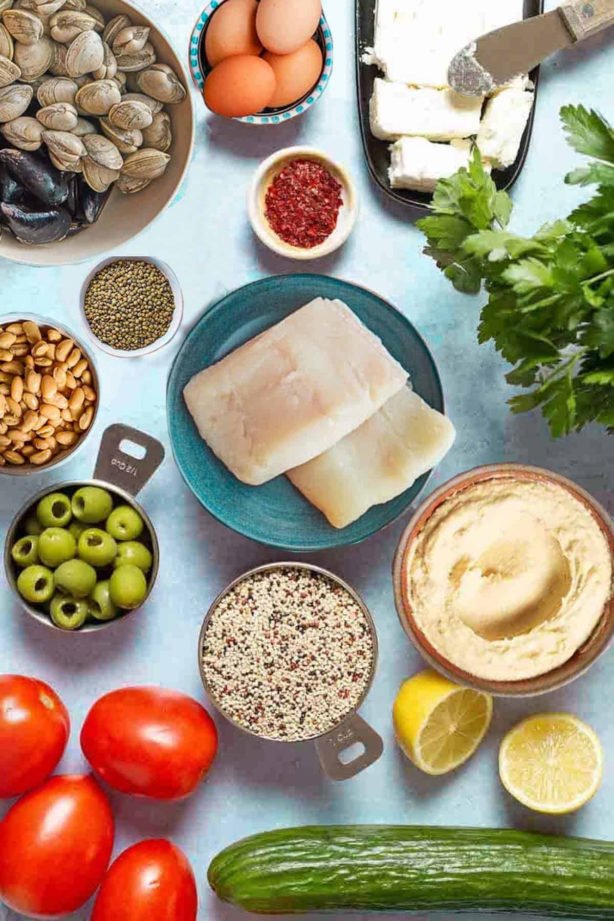 An overhead photo of several Mediterranean diet friend foods including eggs, fish, clams, mussels, feta cheese, aleppo pepper, lentils, quinoa, beans, olives, tomatoes, lemons, cucumber, hummus and parsley.