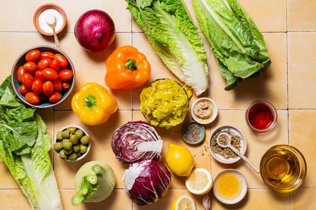 Ingredients for Italian chopped salad including romaine lettuce, radicchio, fennel, red onion, bell peppers, grape tomatoes, green olives, pepperoncini, olive oil, lemon, red wine vinegar, dijon mustard, dried oregano, salt, and black pepper.