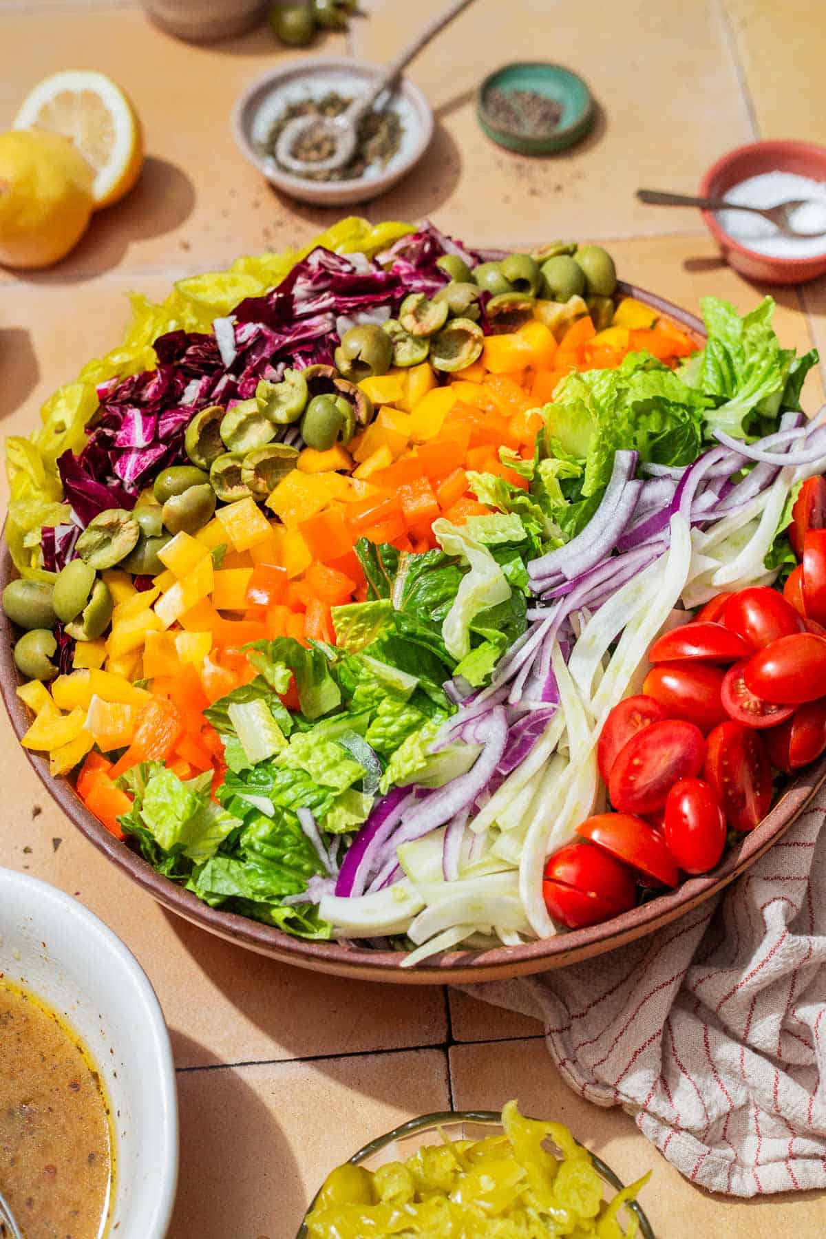 A close up of the ingredients for the chopped salad neatly arranged in a serving bowl, just before being tossed together. Next to this is a bowl of the vinaigrette, lemons, a cloth napkin, and bowls of salt, pepper, vinegrette and pepperoncini.