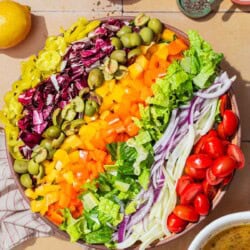 An overhead photo of the ingredients for the chopped salad neatly arranged in a serving bowl, just before being tossed together. Next to this is a bowl of the vinaigrette, lemons, a cloth napkin, and bowls of salt, pepper, and pepperoncini.