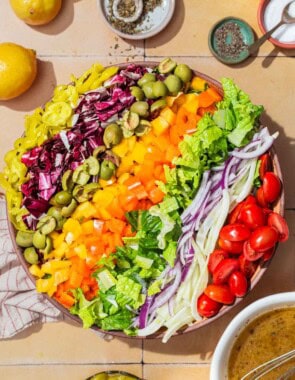 An overhead photo of the ingredients for the chopped salad neatly arranged in a serving bowl, just before being tossed together. Next to this is a bowl of the vinaigrette, lemons, a cloth napkin, and bowls of salt, pepper, and pepperoncini.