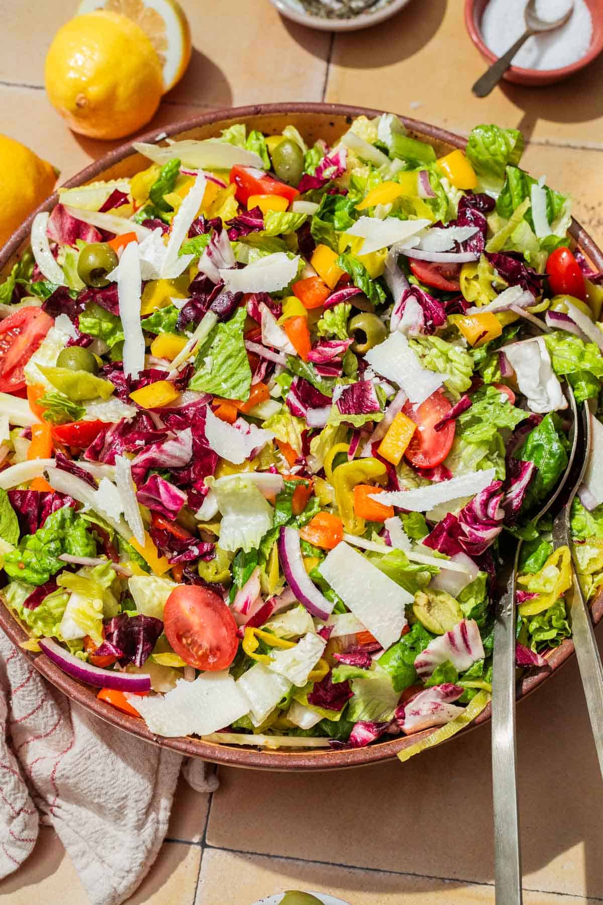 An overhead photo of an Italian chopped salad in a serving bowl with serving utensils. Next to this is a cloth napkin, lemons and a bowl of salt.