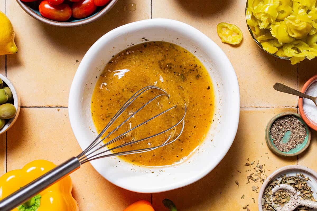 An overhead photo of the vinaigrette for the Italian salad in a bowl with a whisk. Next to this is a lemon, bell peppers, and bowls of salt, pepper, olives, oregano and pepperoncini.