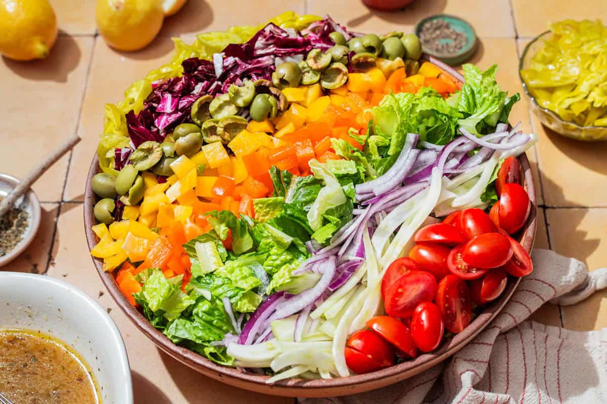 A close up of the ingredients for the chopped salad neatly arranged in a serving bowl, just before being tossed together. Next to this is a bowl of the vinaigrette, lemons, a cloth napkin, and bowls of salt, pepper, and pepperoncini.