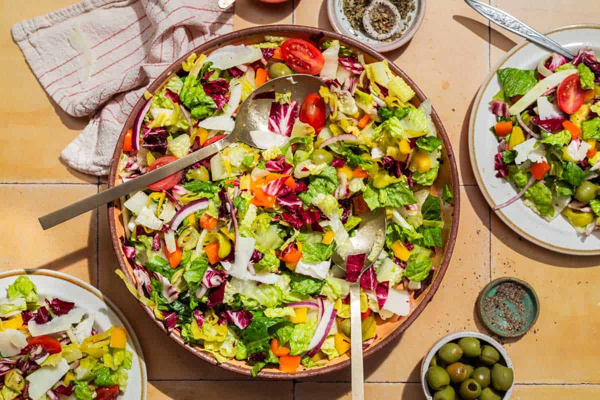 An overhead photo of an Italian chopped salad in a serving bowl with serving utensils. Next to this is a cloth napkin, 2 plates of the salad, and bowls of oregano, black pepper and green olives.