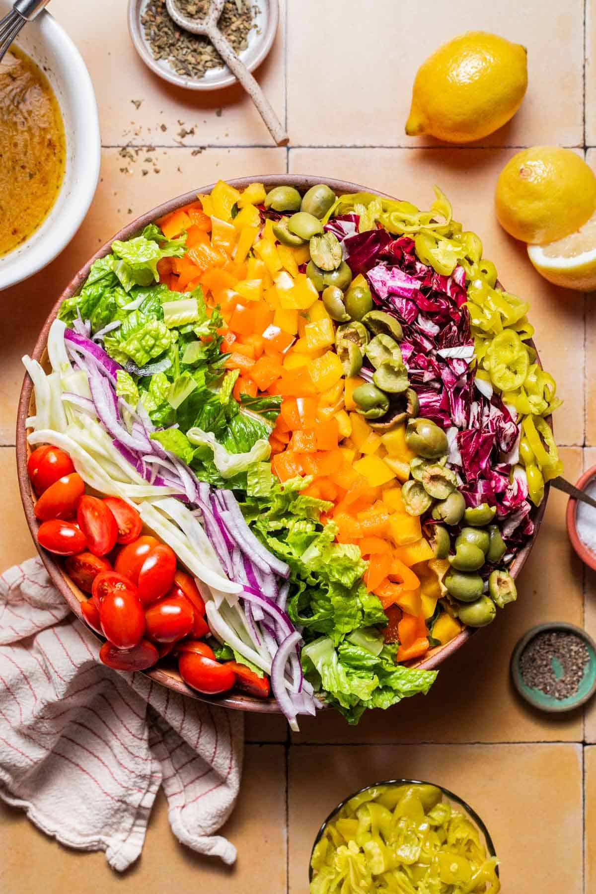 An overhead photo of the ingredients for the chopped salad neatly arranged in a serving bowl, just before being tossed together. Next to this is a bowl of the vinaigrette, lemons, a cloth napkin, and bowls of salt, pepper, oregano and pepperoncini.