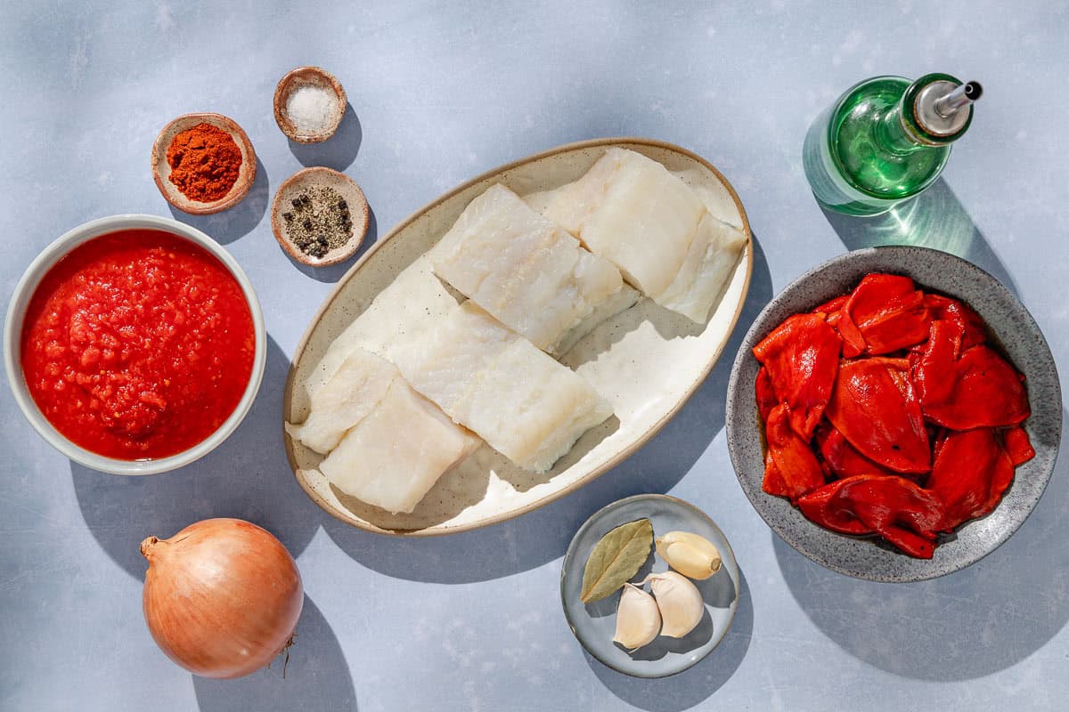 Ingredients for tomato sauce poached cod including cod fillets, olive oil, onion, garlic, roasted peppers, crushed tomatoes, smoked spanish paprika, salt, black pepper, and a bay leaf.