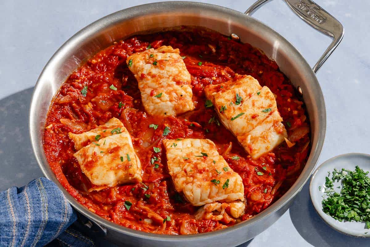 A close up of tomato sauce poached cod fillets in a skillet. Next to this is a bowl of chopped parsley.