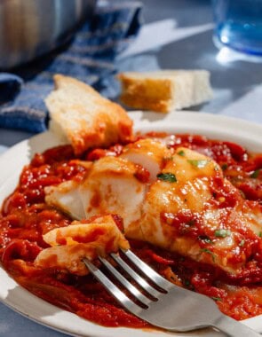 A close up of a tomato sauce poached cod fillet on a plate with a fork and some crusty bread.