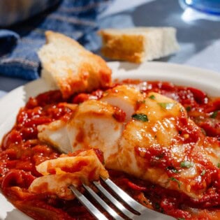 A close up of a tomato sauce poached cod fillet on a plate with a fork and some crusty bread.