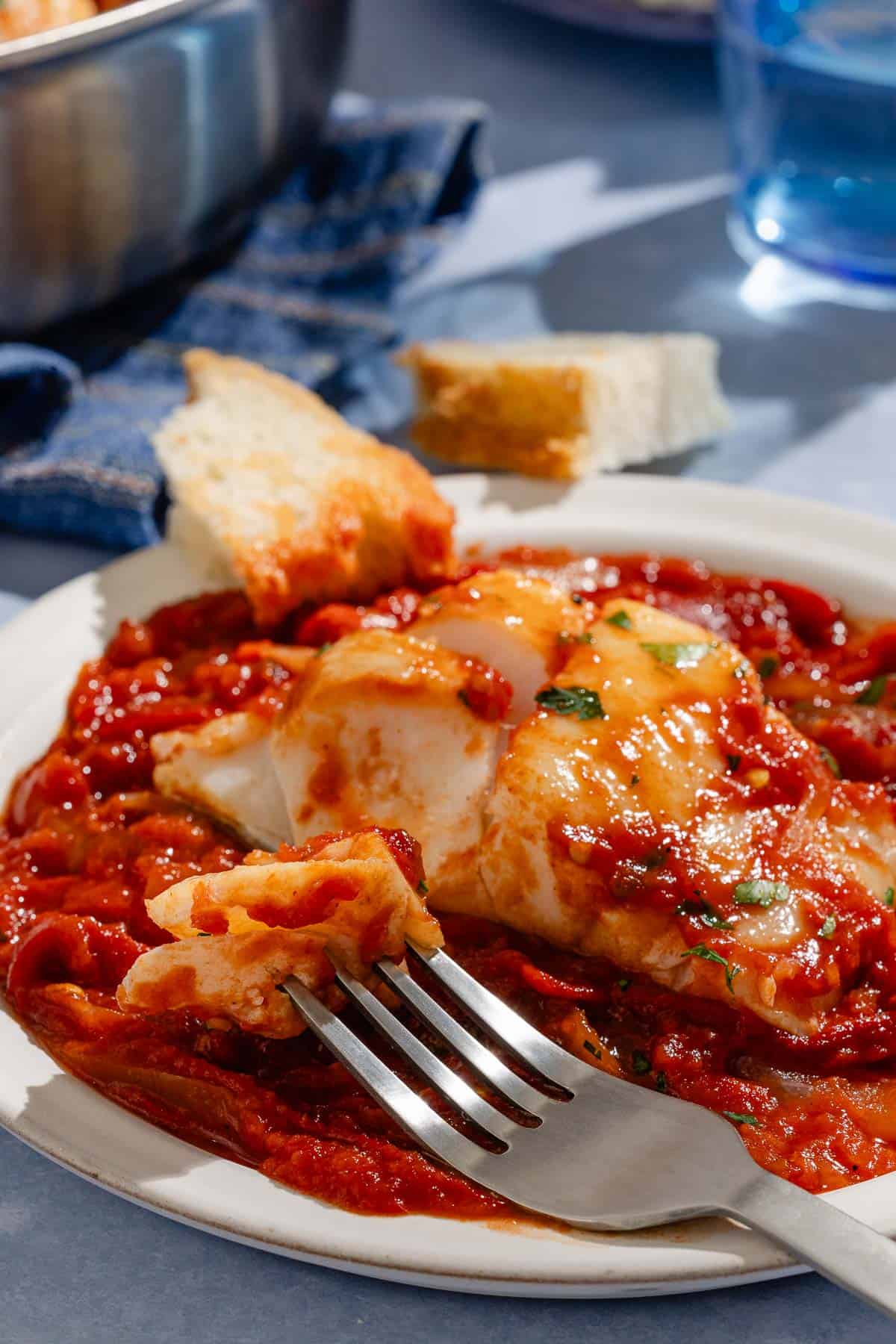 A close up of a tomato sauce poached cod fillet on a plate with a fork and some crusty bread.