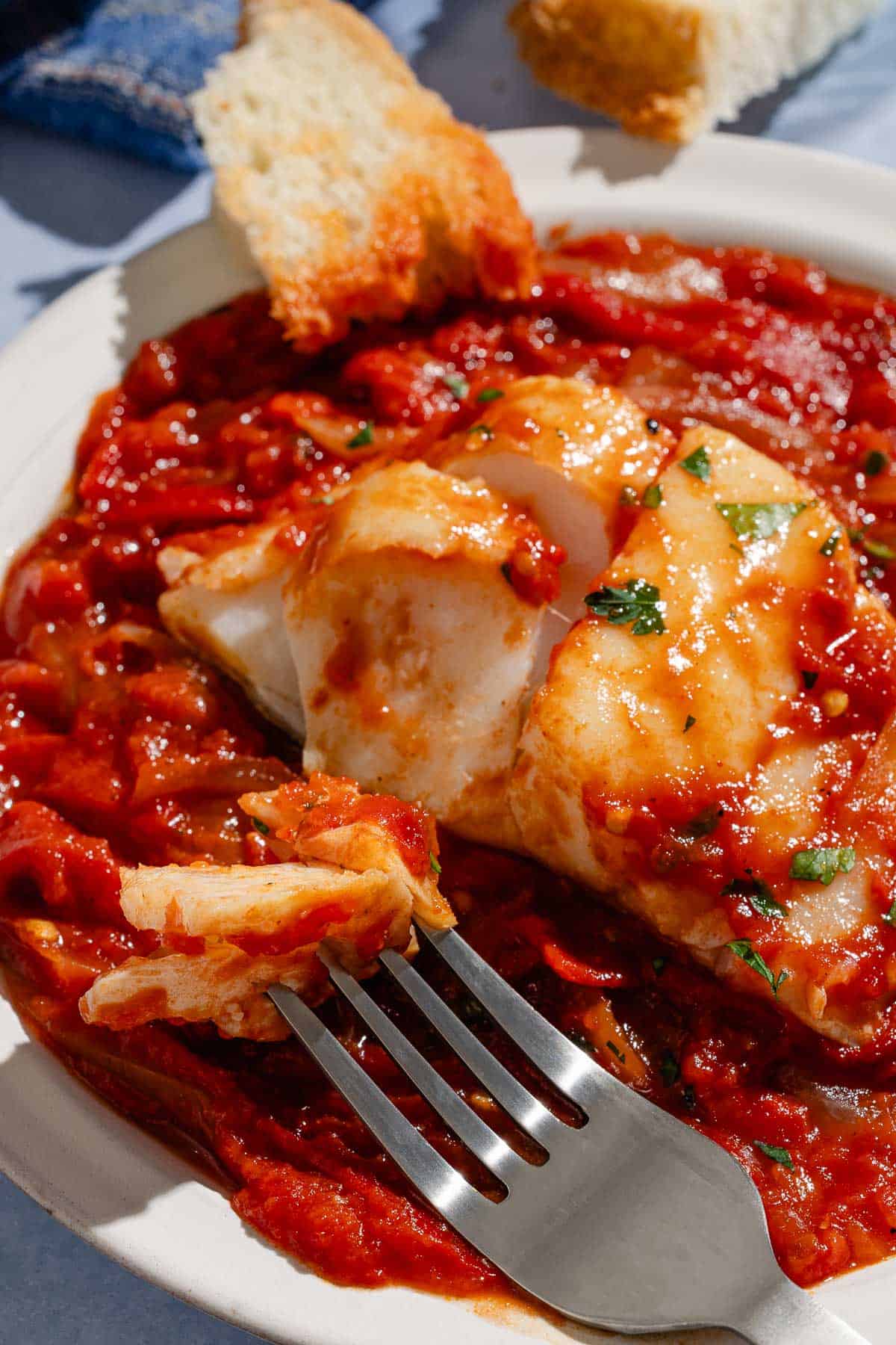 A close up of a tomato sauce poached cod fillet on a plate with a fork and some crusty bread.