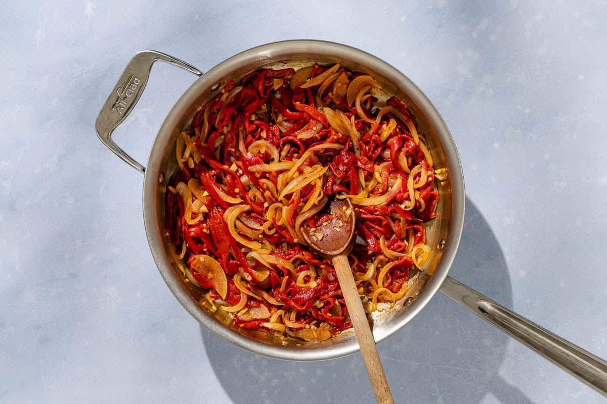 An overhead photo of thinly sliced roasted peppers and onions being sauteed in a skillet with a wooden spoon.