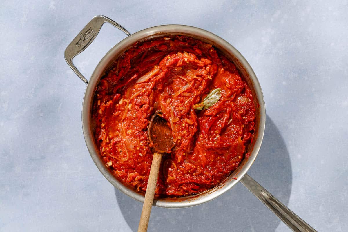 An overhead photo of the tomato sauce for the in a skillet with a bay leaf and a wooden spoo.