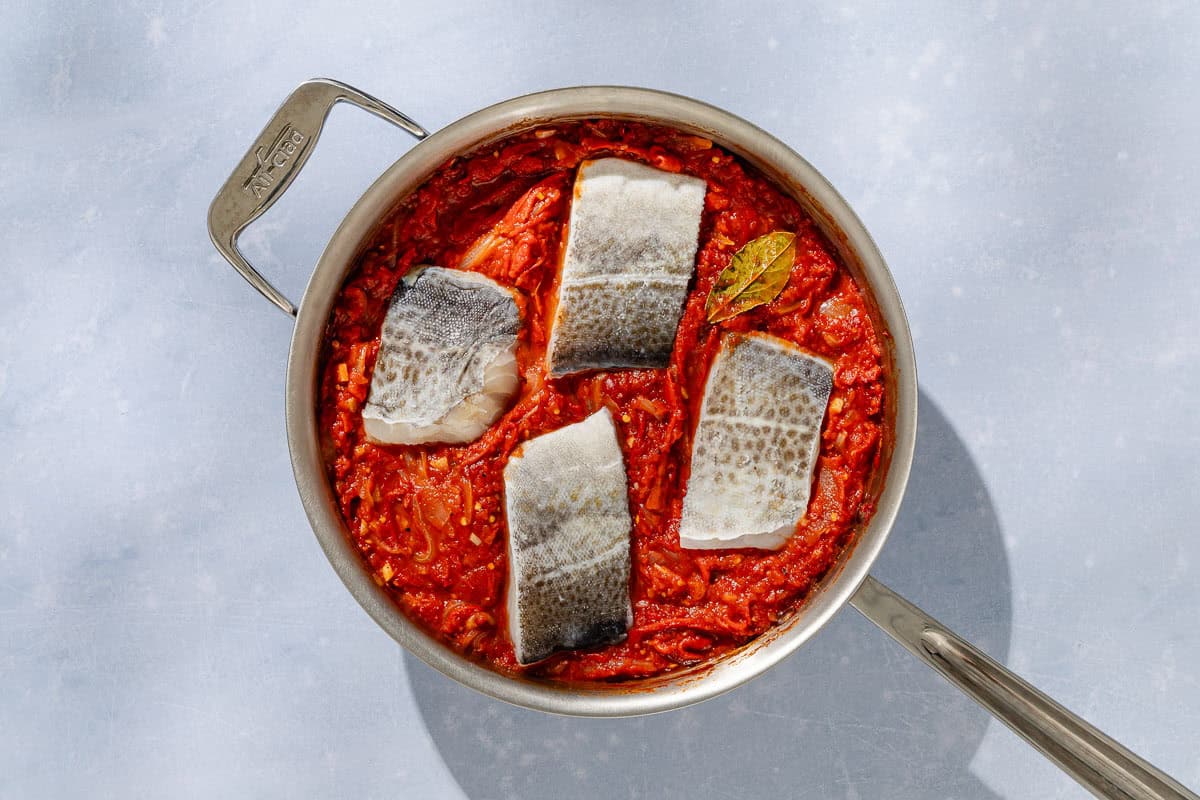 An overhead photo of 4 cod fillets being poached skin up in tomato sauce in a skillet.