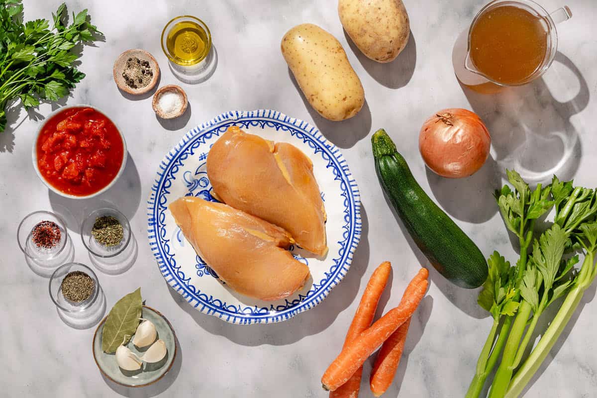 Ingredients for chicken vegetable soup including chicken breasts, carrots, celery, onion, garlic, potatoes, zucchini, a bay leaf, parsley, olive oil, salt, black pepper, dried thyme, oregano, red pepper flakes, caned diced tomatoes, and chicken stock.