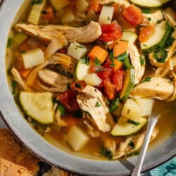 An overhead photo of a serving of chicken vegetable soup in a bowl with a spoon next to pieces of crusty bread.