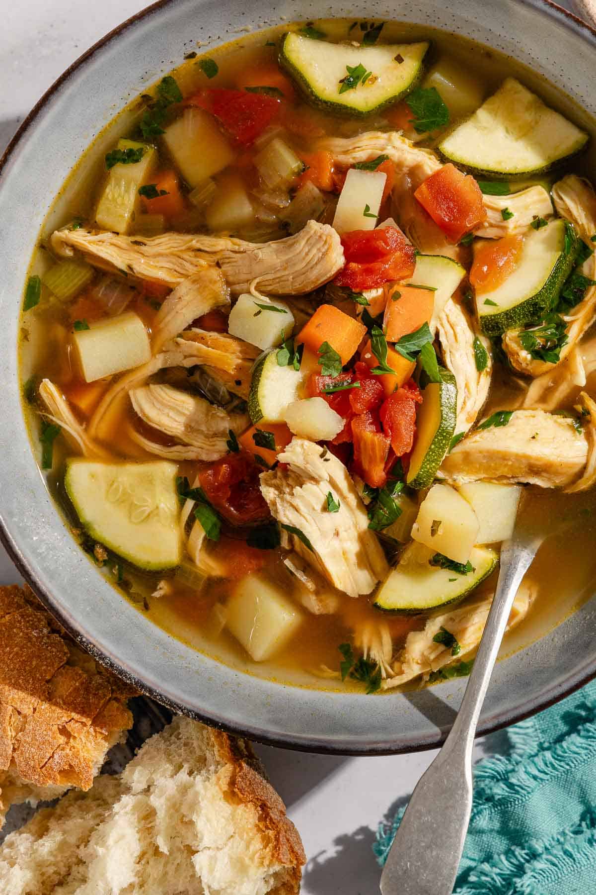 An overhead photo of a serving of chicken vegetable soup in a bowl with a spoon next to pieces of crusty bread.