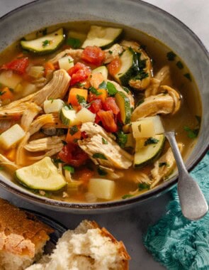 A close up of a serving of chicken vegetable soup in a bowl with a spoon next to pieces of crusty bread.