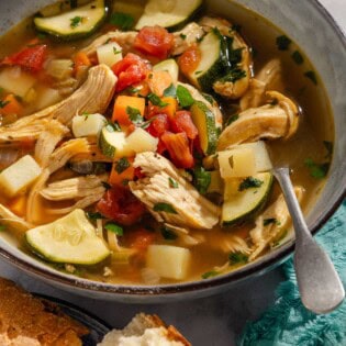 A close up of a serving of chicken vegetable soup in a bowl with a spoon next to pieces of crusty bread.