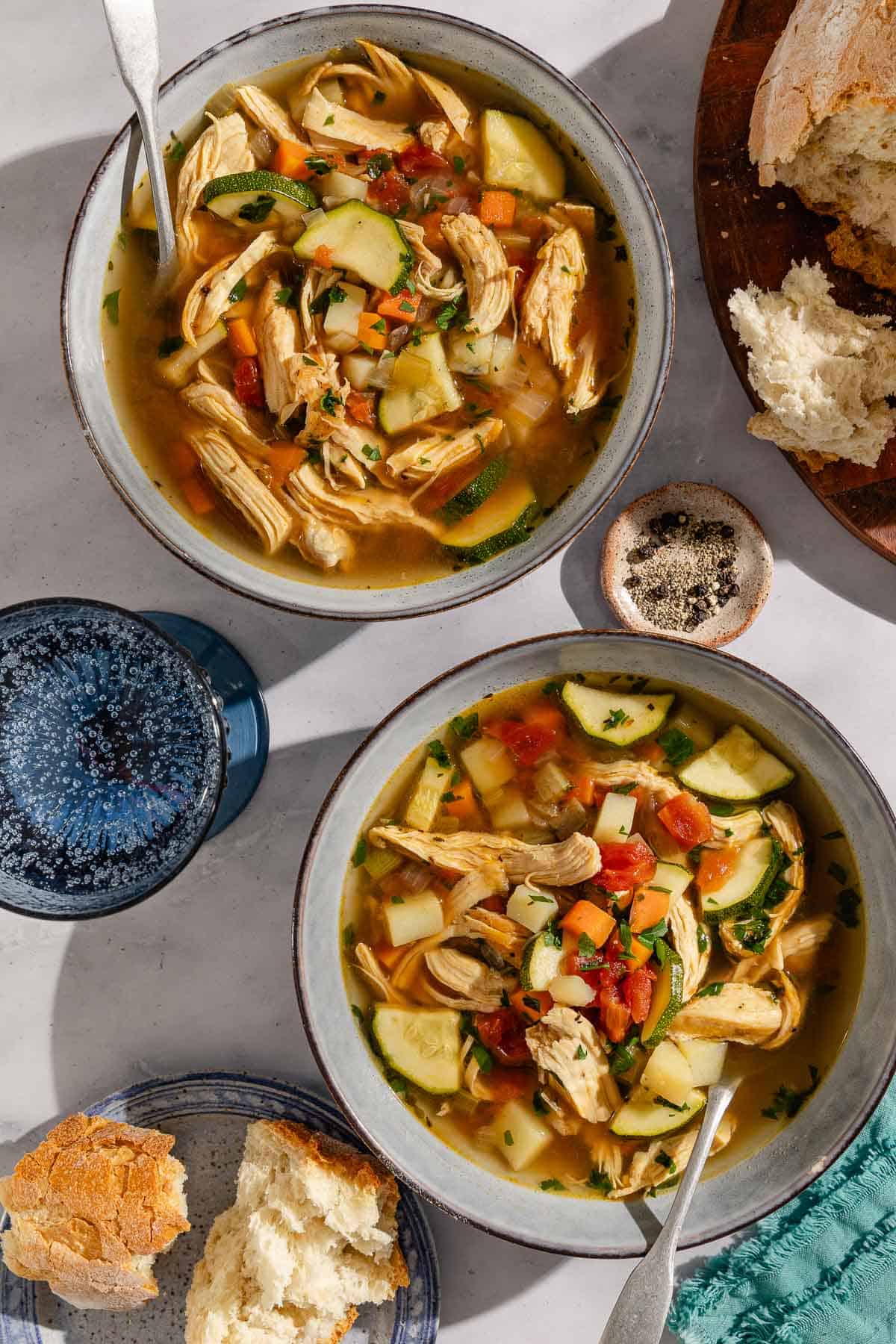 An overhead photo of 2 bowls chicken vegetable soup in bowls with spoons. Next to these is a bowl of black pepper, a glass of sparkling water, plates of crusty bread and a cloth napkin.