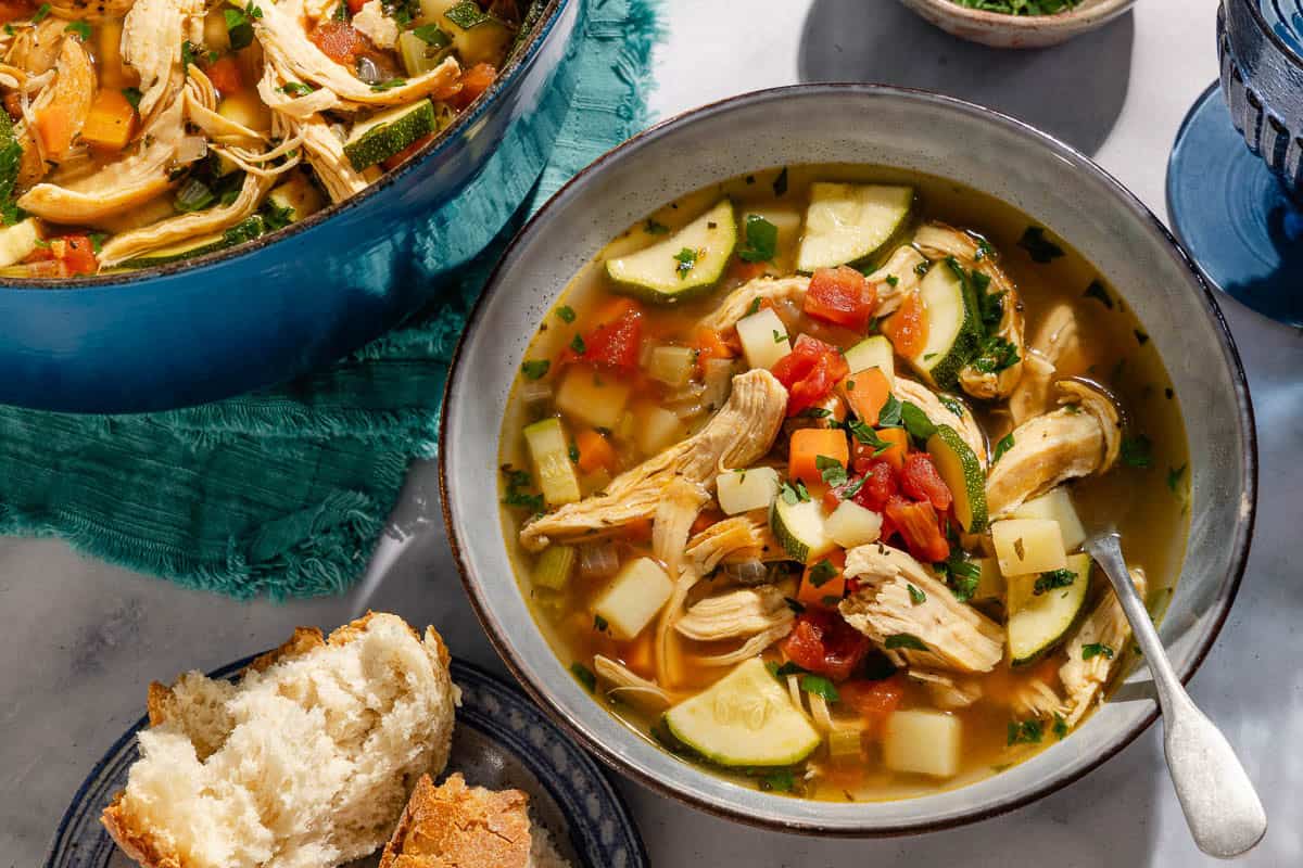 A close up of a serving of chicken vegetable soup in a bowl with a spoon. Next to this is the rest of the soup in a dutch oven and pieces of crusty bread on a plate.