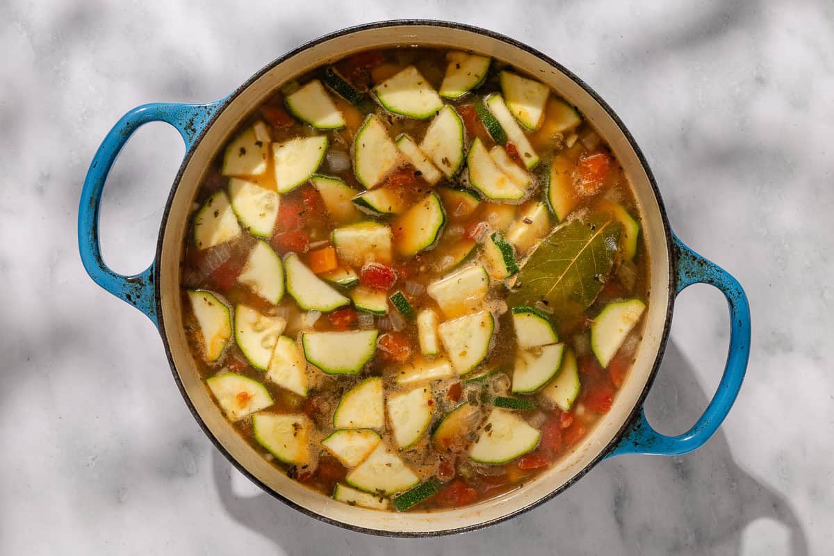 An overhead photo of the vegetables and canned tomatoes for the chicken vegetable soup simmering in chicken broth in a dutch oven with a bay leaf.