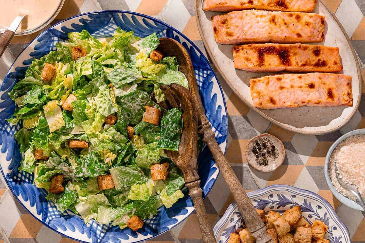 An overhead photo of caesar salad in a serving bowl with wooden serving utensils. Next to this is a plate of cooked salmon filets, and bowls of croutons, caesar salad dressing, grated parmesan cheese and black pepper.