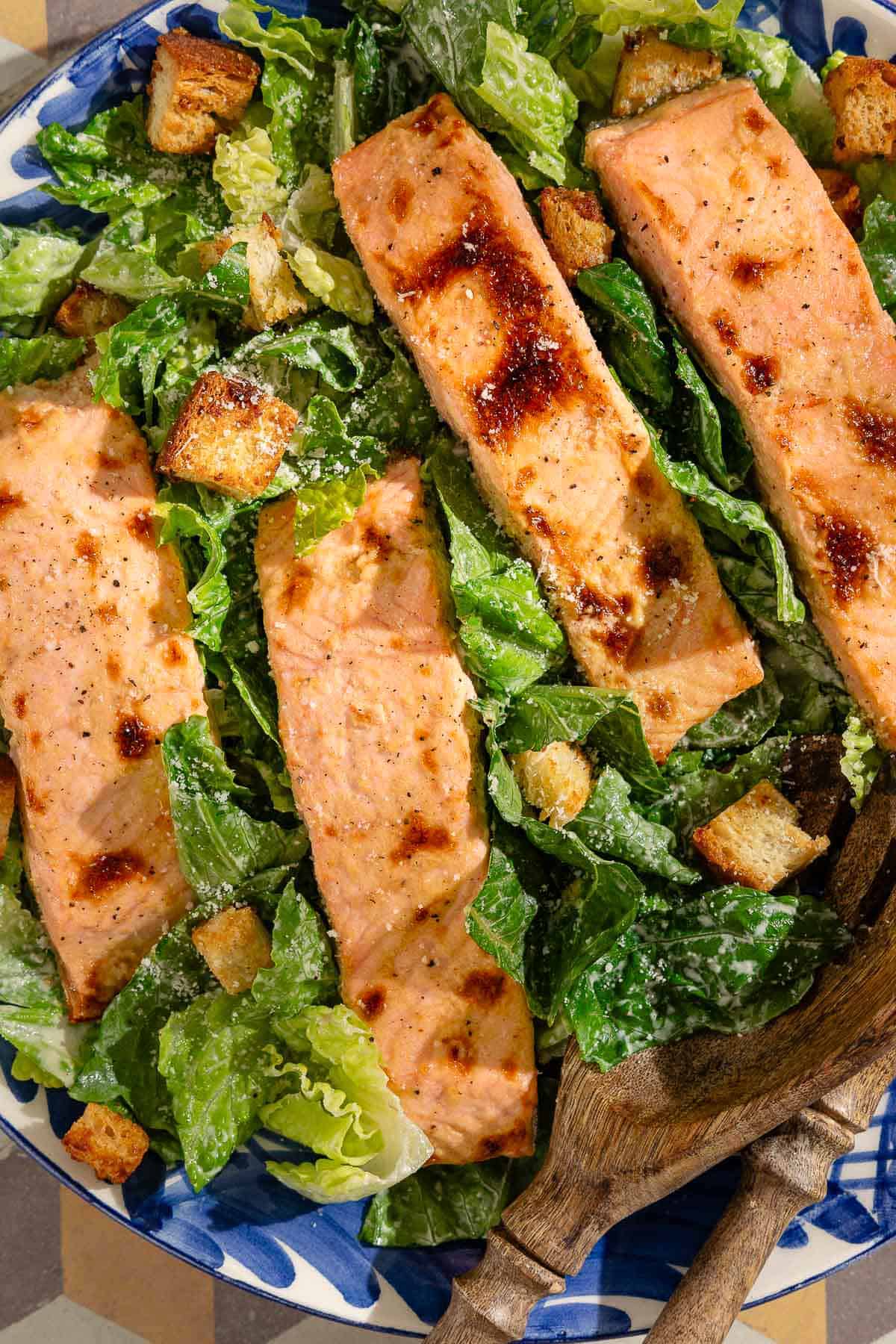 A close up of salmon caesar salad in a serving bowl with wooden serving utensils.