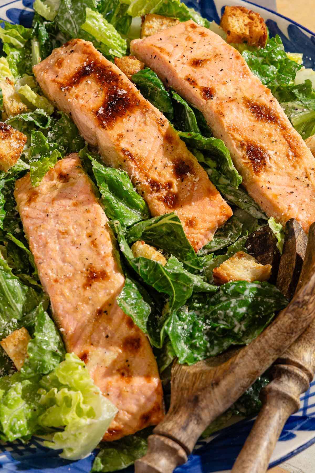 A close up of salmon caesar salad in a serving bowl with wooden serving utensils.