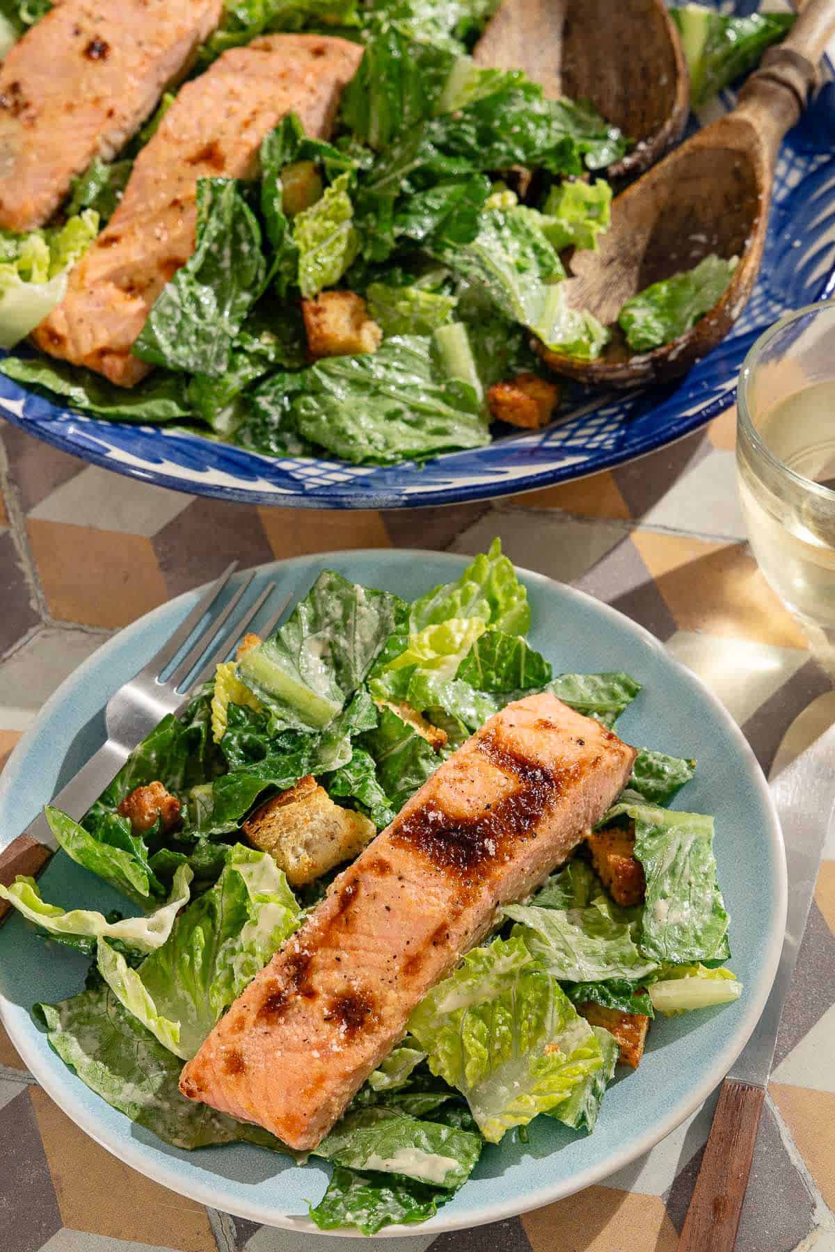 A serving of the salmon caesar salad in a bowl with a fork. Next to this is the rest of the salmon caesar salad in a serving bowl with wooden serving utensils.