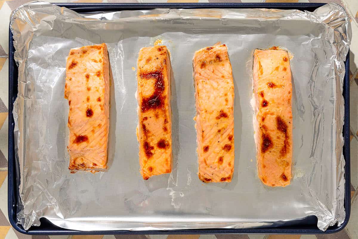 An overhead photo of 4 marinated cooked salmon filets on an aluminum foil lined sheet pan.