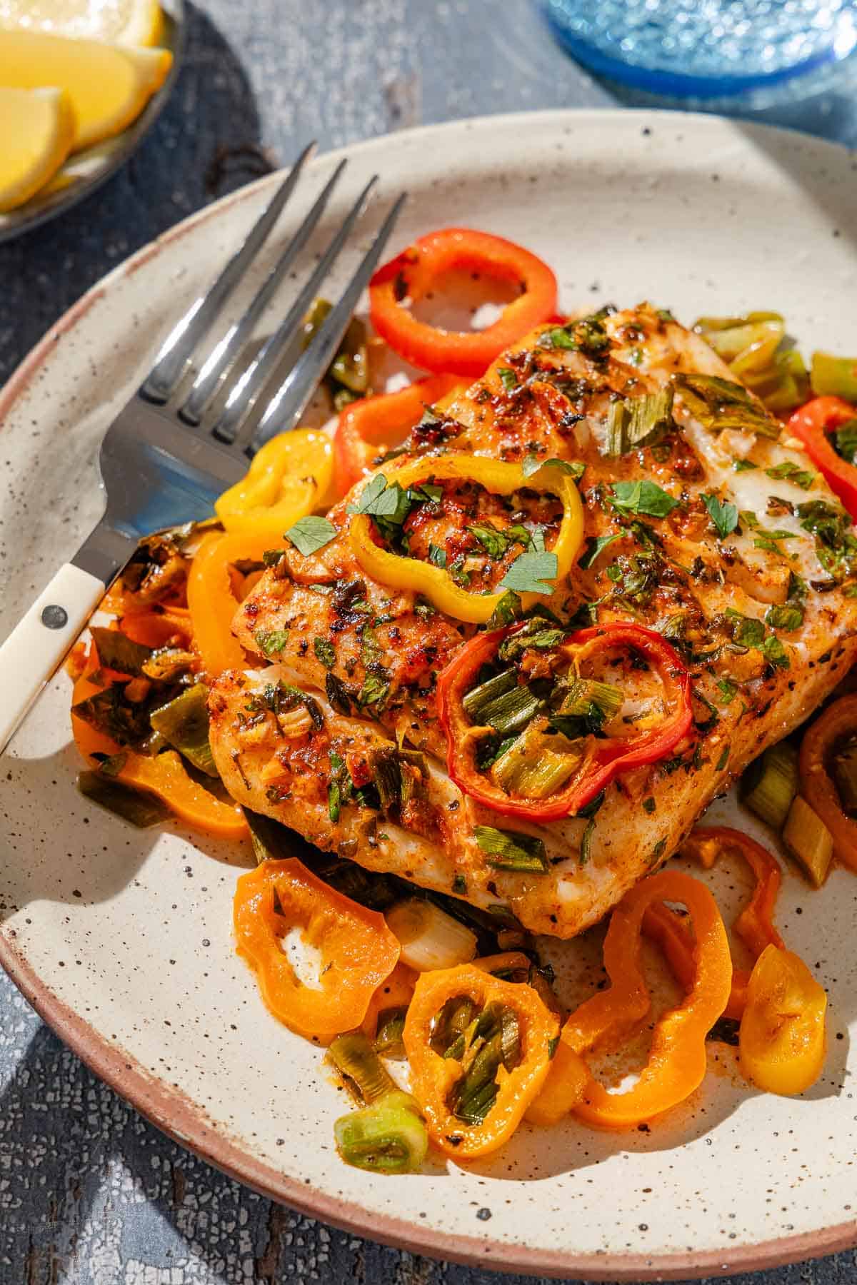 A close up of a serving of baked haddock with baby bell peppers on a plate with a fork.