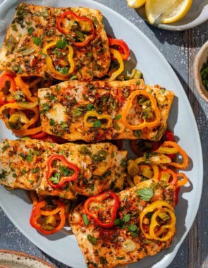 An overhead photo of baked haddock with baby bell peppers on a platter. Next to this is a plate of lemon wedges.