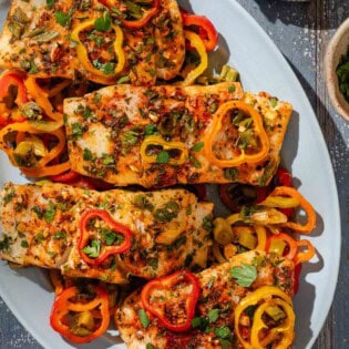 An overhead photo of baked haddock with baby bell peppers on a platter. Next to this is a plate of lemon wedges.