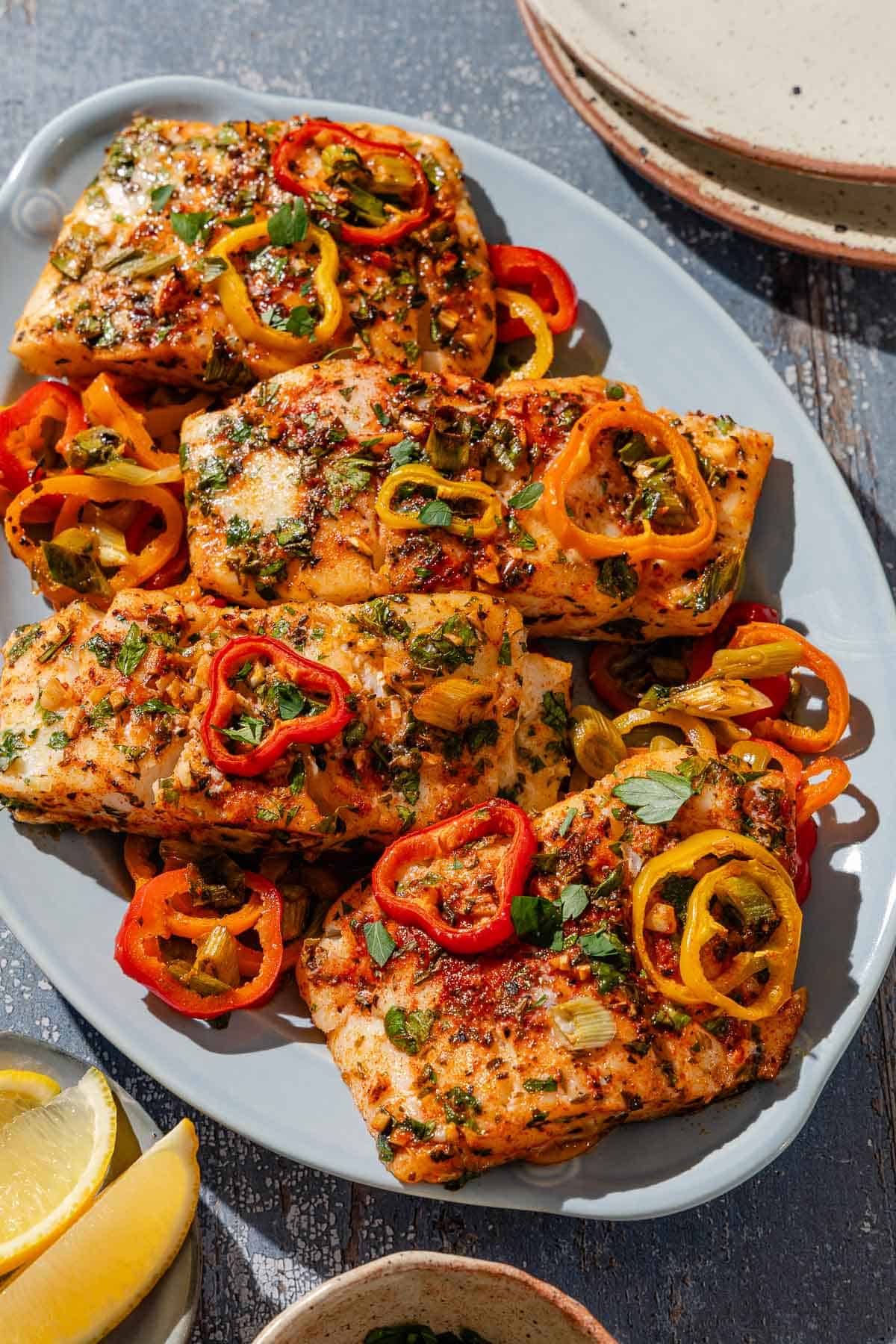 An overhead photo of baked haddock with baby bell peppers on a platter. Next to this is a plate of lemon wedges and a stack of 2 plates.