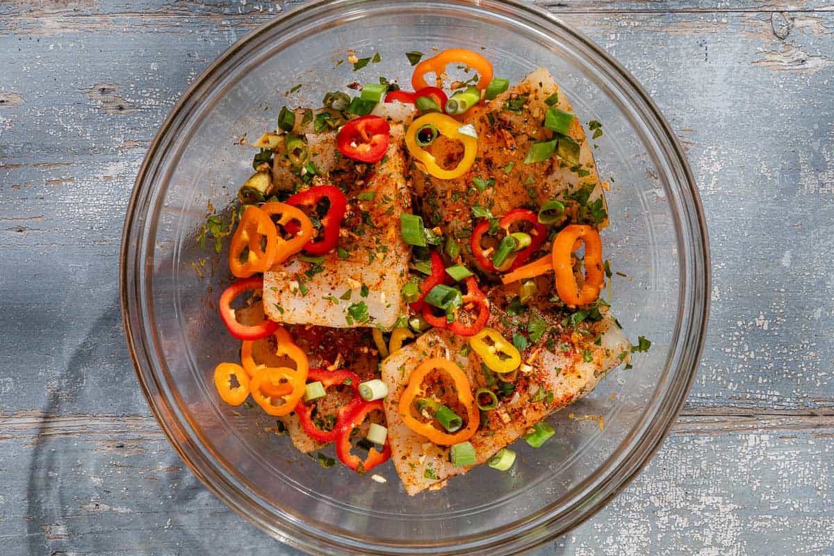 An overhead photo of the seasoned uncooked haddock in a bowl with the baby bell peppers, green onions, garlic and parsley in a bowl.