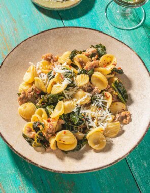 Orecchiette with sausage and broccoli rabe on a plate. Next to this is a glass of white wine and a small bowl of grated pecorino romano cheese.