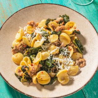 Orecchiette with sausage and broccoli rabe on a plate. Next to this is a glass of white wine and a small bowl of grated pecorino romano cheese.