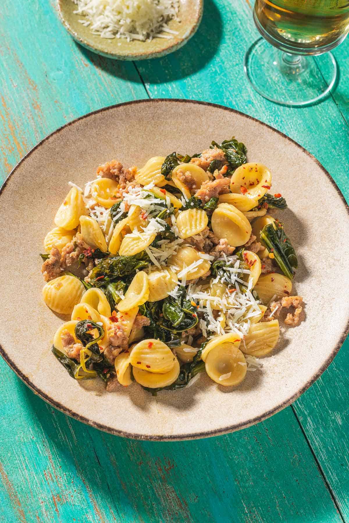 Orecchiette with sausage and broccoli rabe on a plate. Next to this is a glass of white wine and a small bowl of grated pecorino romano cheese.