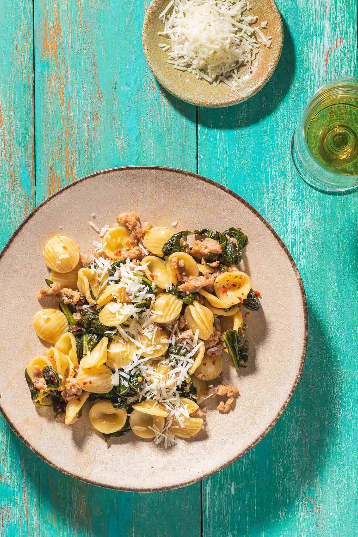 An overhead photo of orecchiette with sausage and broccoli rabe on a plate. Next to this is a glass of white wine and a small bowl of grated pecorino romano cheese.