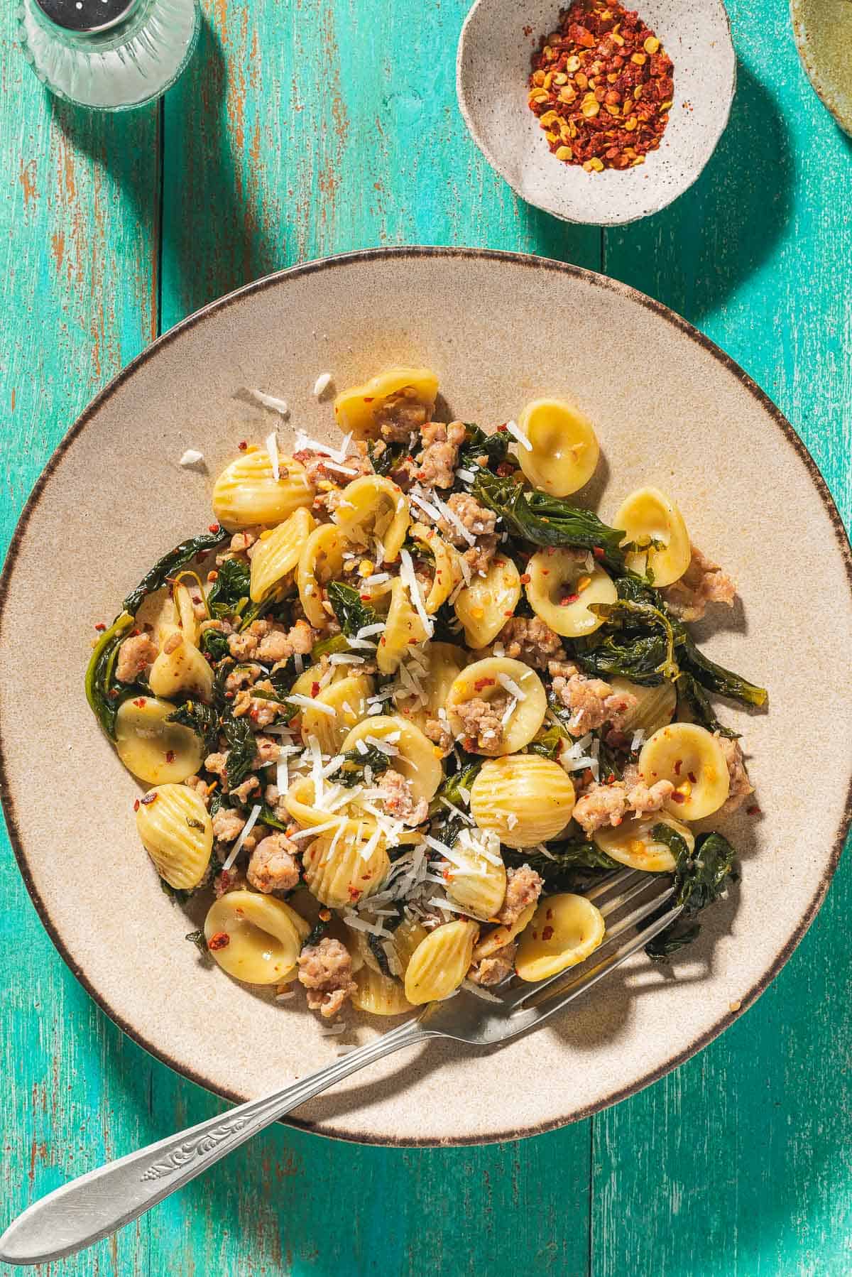 An overhead photo of orecchiette with sausage and broccoli rabe on a plate with a fork. Next to this is a salt shaker and a small bowl of red pepper flakes.