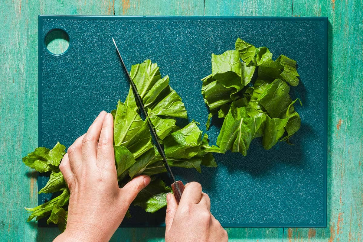 An overhead photo of the stalks of broccoli rabe being chopped with a knife on a cutting board.