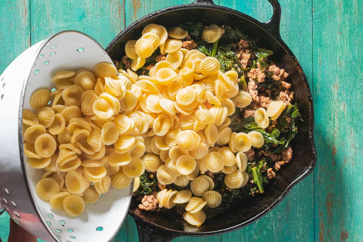 An overhead photo of cooked orecchiette pasta being poured from a colander into the pan with the broccoli rabe and sausage.