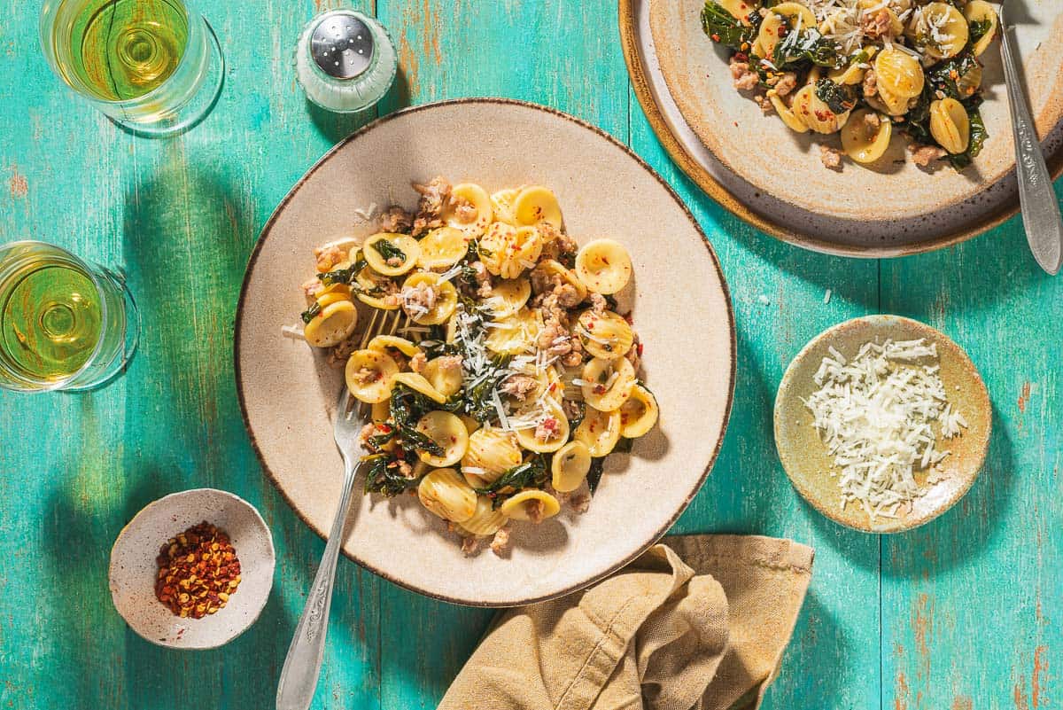 An overhead photo of orecchiette with sausage and broccoli rabe on a plate with a fork. Next to this is another plate of the orecchiette, a salt shaker, 2 glasses of white wine, a cloth napkin,and small bowls of red pepper flakes and grated pecorino romano cheese.