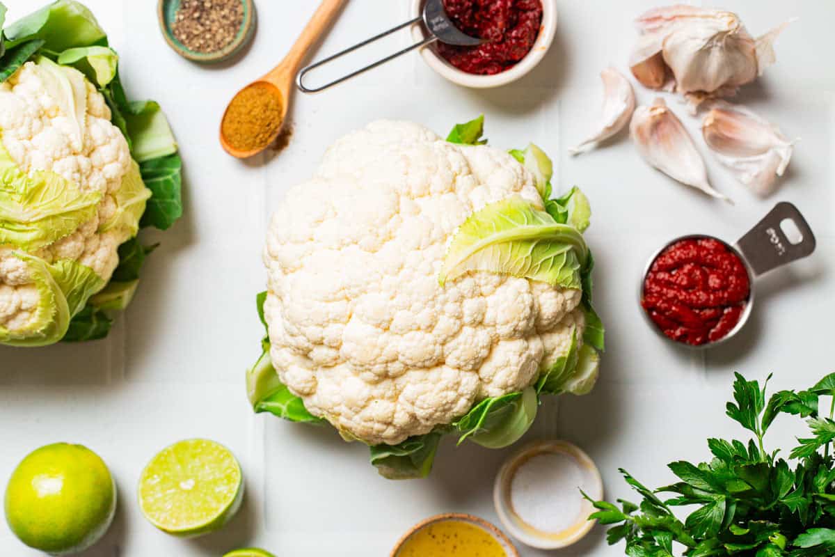 Ingredients for spicy baked cauliflower including 2 heads of cauliflower, garlic, parsley, ground cumin, harissa, tomato paste, limes, olive oil, salt and black pepper.