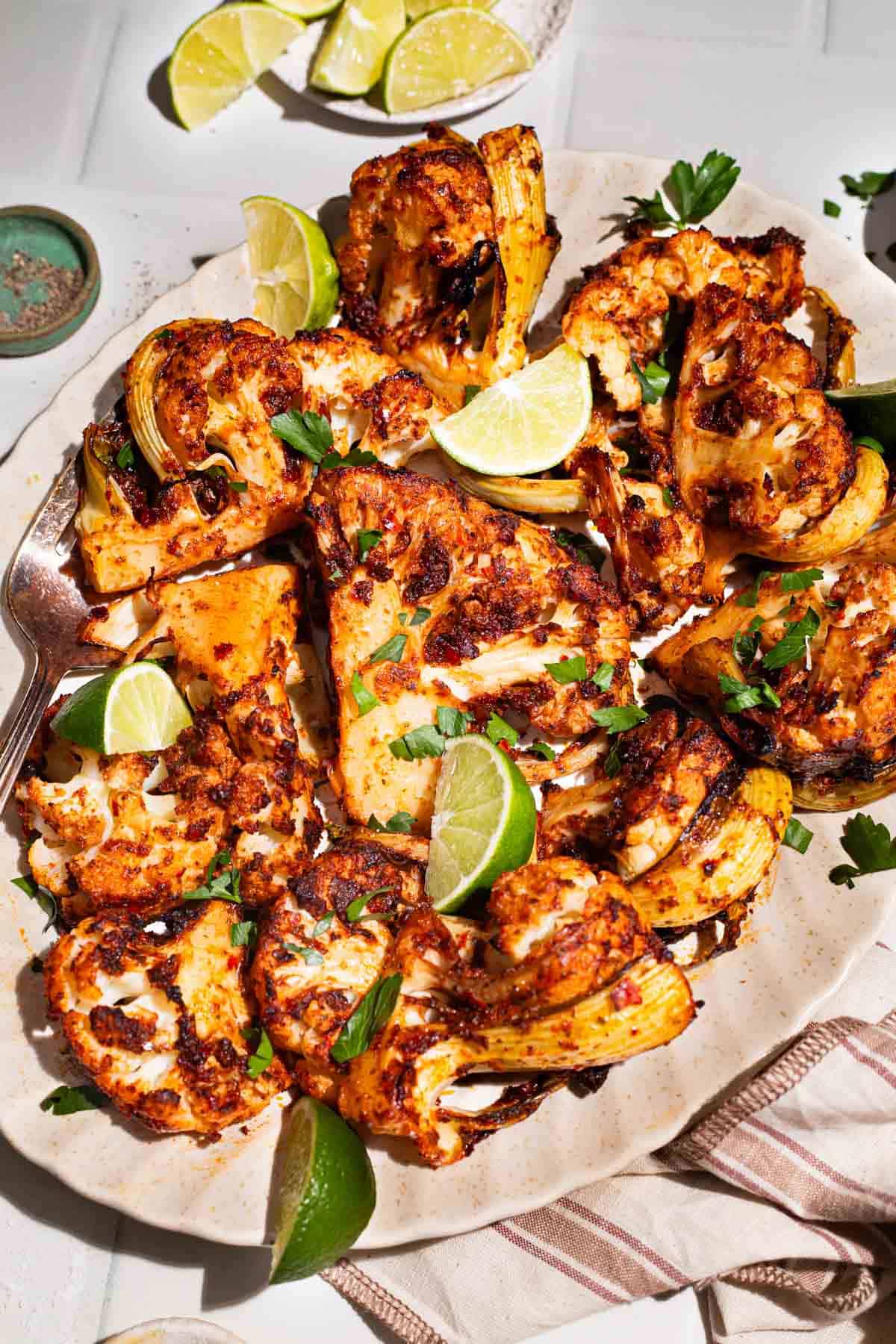 An overhead photo of spicy baked cauliflower topped with chopped parsley and lime wedges on a serving platter with a fork. Next to this is a plate of lime wedges, a cloth napkin, and a bowl of black pepper.